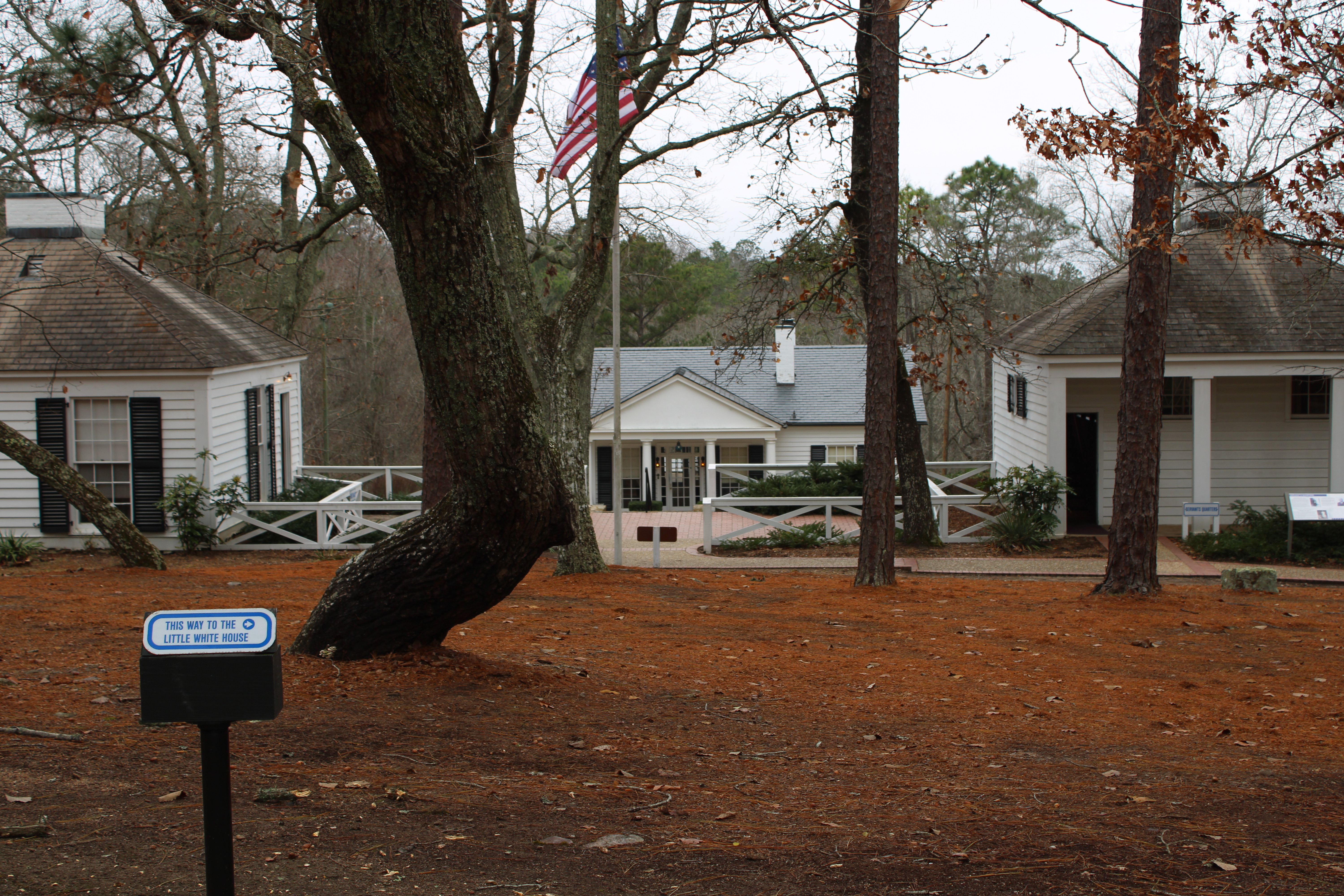 A view of the main cottage, alongside the servants quarters on the right and guest house on the left. Large trees block some of the view of the main cottage. The American flag is also seen in front of the main cottage